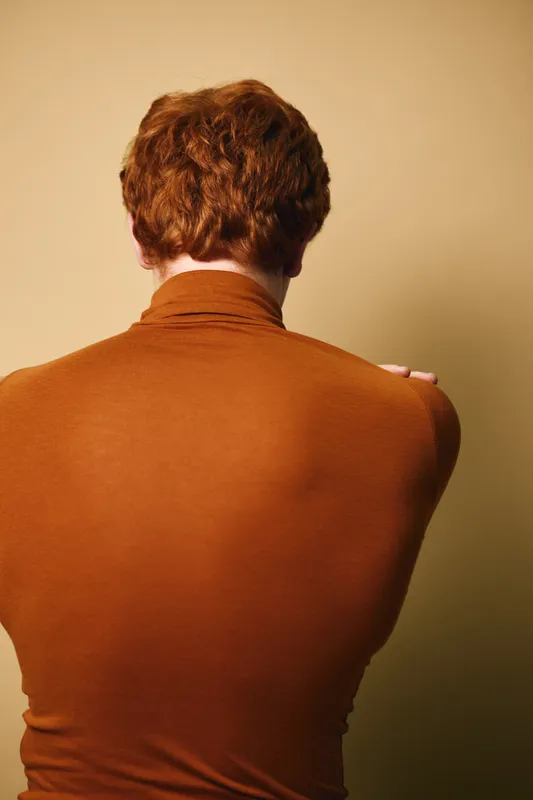 Man with red hair wearing brown turtleneck sweater posing with back to camera on beige background. Casual style, hairstyle and upper body in warm tones for fashion or portrait concept.