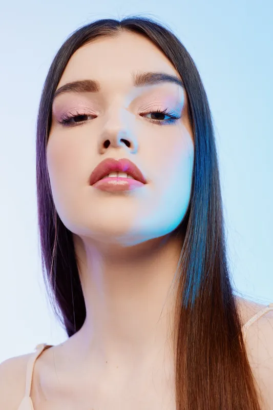Young woman with long hair poses in a soft blue studio light. Her makeup is subtle with pink lips and light eyeshadow. Concept of beauty, fashion, and elegance.