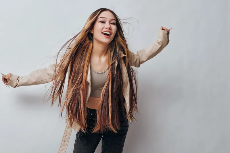 Young woman with long braided hair and casual clothes smiling joyfully, posing with arms raised on plain gray background for lifestyle and happiness concept.