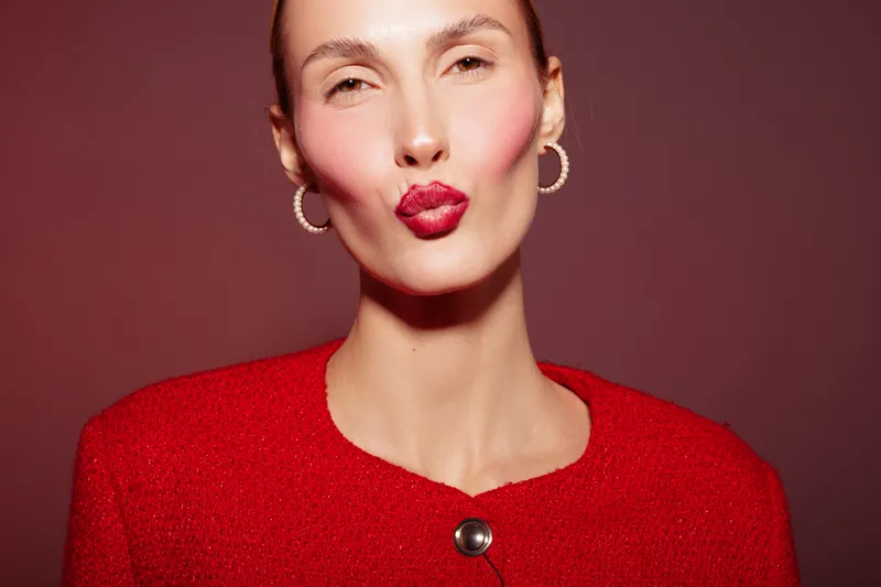 Close-up of a woman with red lipstick and red jacket blowing a kiss on a red background. Concept of beauty, fashion, makeup, and romantic expression in studio lighting.