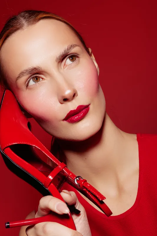 Close-up of a woman with glossy red lips holding a red high heel shoe near her face. The red background and outfit create a bold, stylish, and fashion-forward concept.