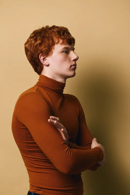 Young man with red hair wearing brown turtleneck sweater stands sideways with arms crossed against beige background. Profile portrait with serious expression and casual style.