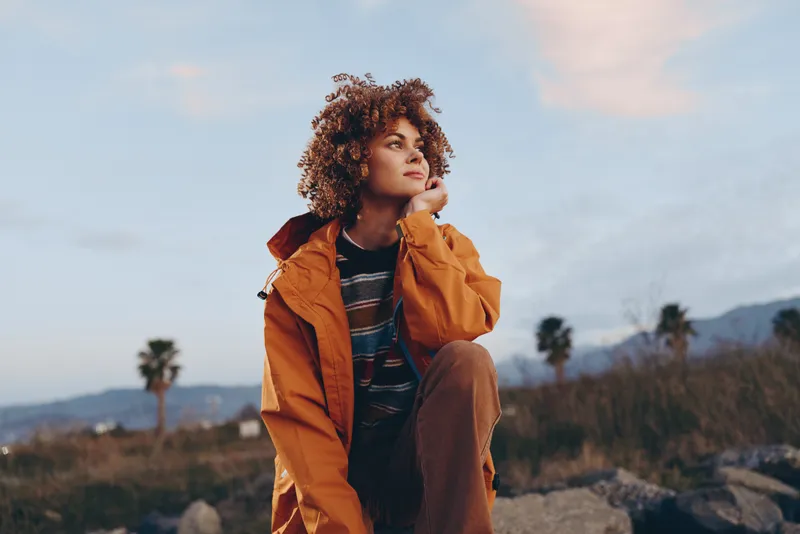 Woman lifestyle outdoors in an orange jacket, curly hair framing her face, thoughtful pose at sunset, soft breeze, warm natural light, scenic landscape with distant trees, today