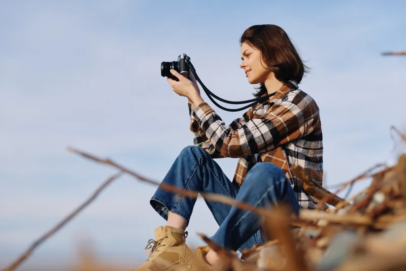 Woman photographer sits among dry branches, holding a camera for outdoor nature photography in a casual plaid shirt and jeans, capturing a peaceful landscape with focused expression.