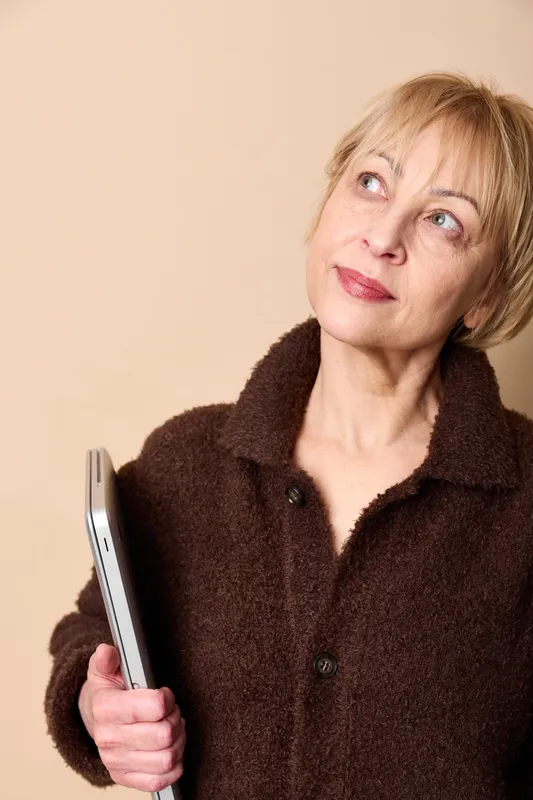 Woman portrait, mature blonde in indoor setting, holding a smartphone, thoughtful gaze upward, soft light, beige studio background, casual coat, calm expression, closeup