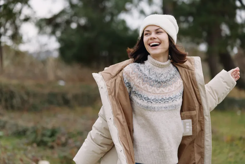 Woman enjoying outdoor weather in a warm sweater and beanie, wearing a light jacket, smiling, in a park setting with trees and open space, autumn mood and relaxed casual style.