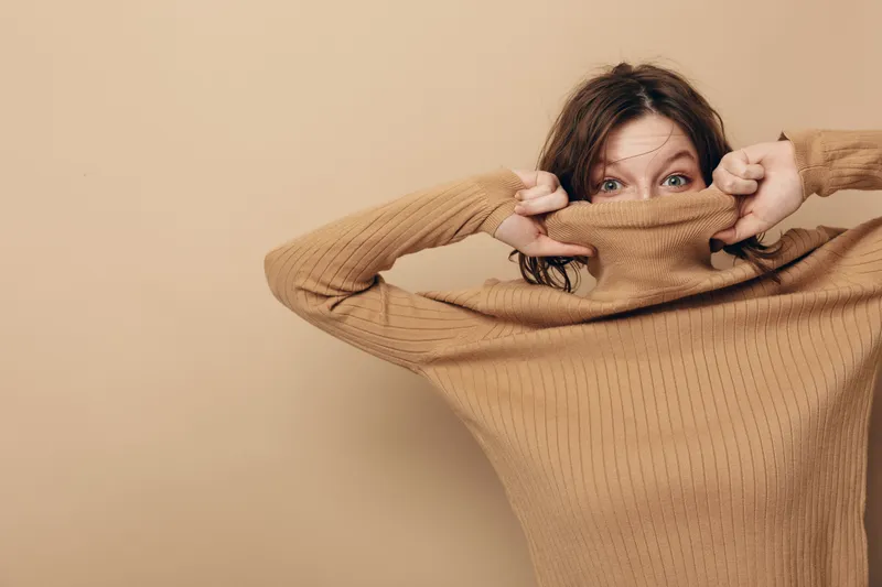 Young woman in brown knitted sweater pulls collar over face, peeking with blue eyes. Studio portrait on beige background. Concept of cozy fashion, autumn style, and playful mood.