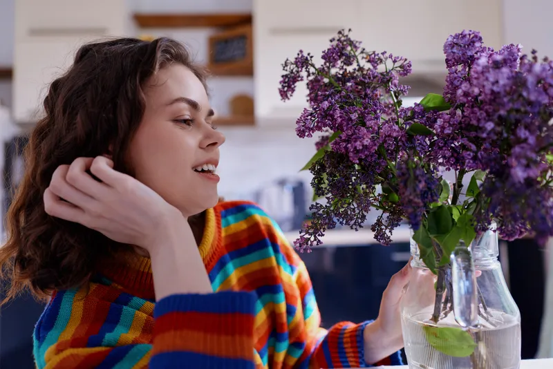 A woman in a bright striped sweater sits at home, admiring a vase of purple flowers on the kitchen counter, capturing a cozy, relaxed lifestyle moment of everyday care in a warmly lit, inviting scene.