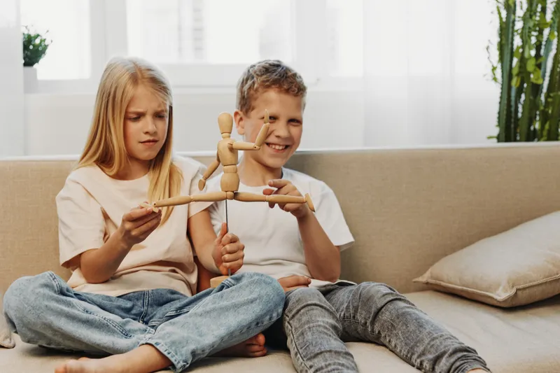 Two children sitting on sofa play with wooden mannequin toy, showing curiosity and joy in bright living room. Concept of childhood, creativity, and sibling interaction.