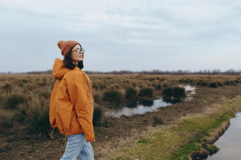 Woman lifestyle outdoors in a rural landscape, wearing an orange jacket and knit hat, walking by a small stream in a grassy field under cloudy sky, calm nature mood, soft natural light