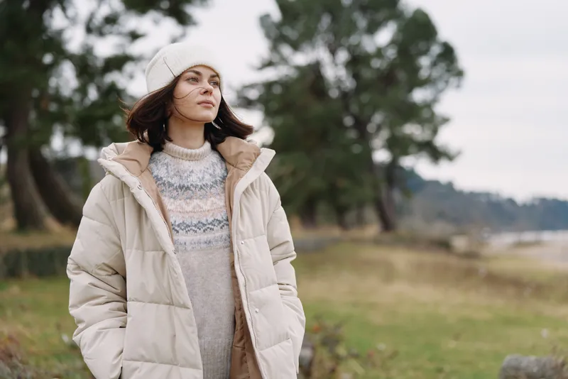 Woman, outdoor, winter, coat, beanie, landscape, field, calm in a beige puffer on a breezy day, looking ahead with a serene mood amid open countryside and trees
