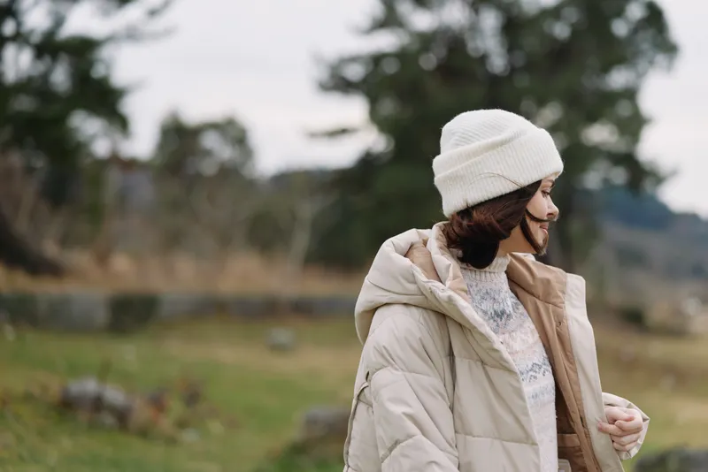 Woman in a puffy coat and white beanie stands in a windy outdoor landscape near a lake with trees and hills behind, conveying a calm moment