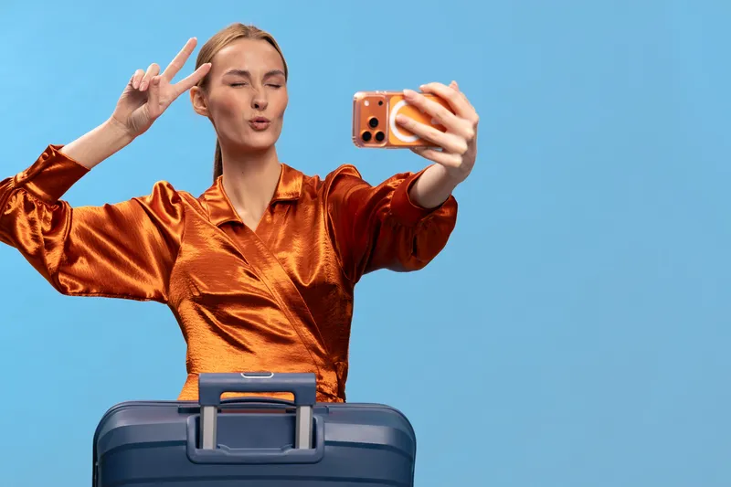 Young woman in orange shiny blouse takes selfie with peace sign while standing behind blue suitcase on blue background. Concept of travel, vacation, and social media content creation.