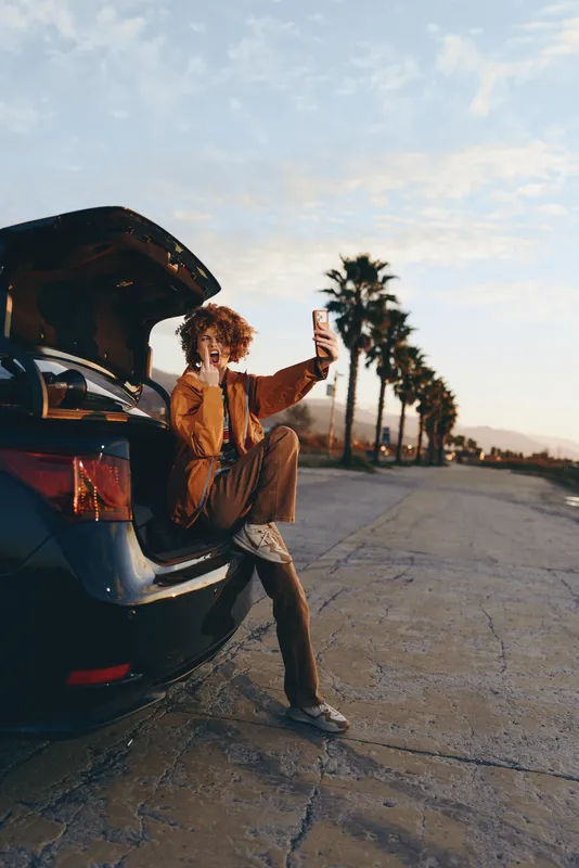 Woman lifestyle, sitting by open car on outdoor road, taking selfie with smartphone at sunset, curly hair in warm breeze, casual outfit, palm lined desert highway, natural light