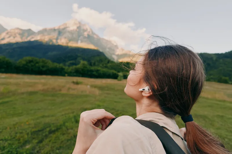 A young woman with brunette hair stands in a grassy field with a backpack, gazing toward distant mountains under a bright sky, conveying a moment of calm and exploration.