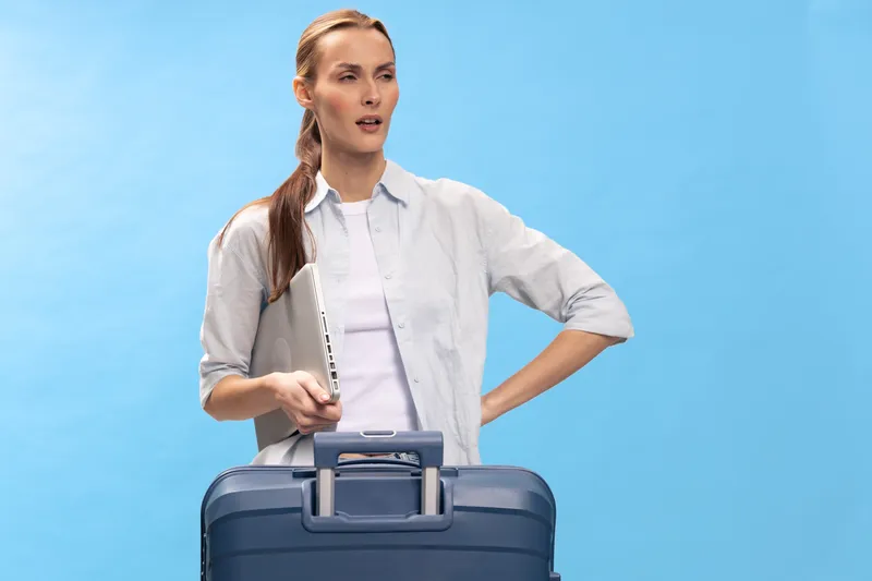 Young woman with suitcase holds laptop and looks thoughtful on blue background. Concept of travel planning, business trip, and decision making with casual outfit and natural light.