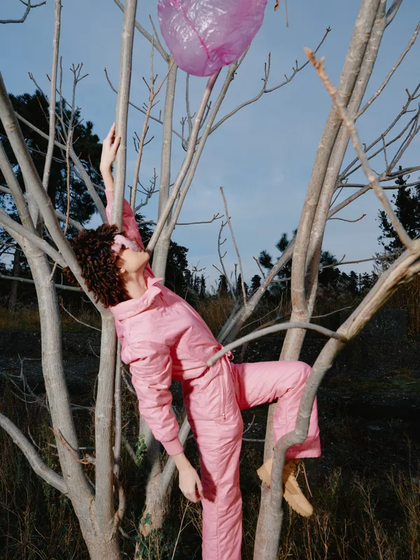 Young person climbing a bare tree in a pink jumpsuit reaching for a pink balloon outdoors, in an open field with blue sky and distant trees, a playful moment of ascent and curiosity.
