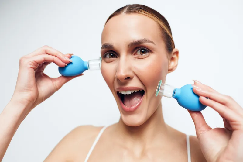 Cheerful woman performing ear care at home, smiling with mouth open, holding blue bulb devices near ears, bright studio light, casual setting, healthy lifestyle