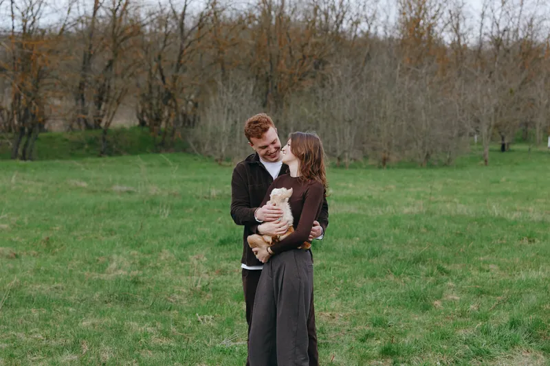 A couple outdoors in a grassy park, smiling as they cradle a young goat kid between them. Bright natural light, casual attire, candid lifestyle moment capturing affection, companionship, and gentle