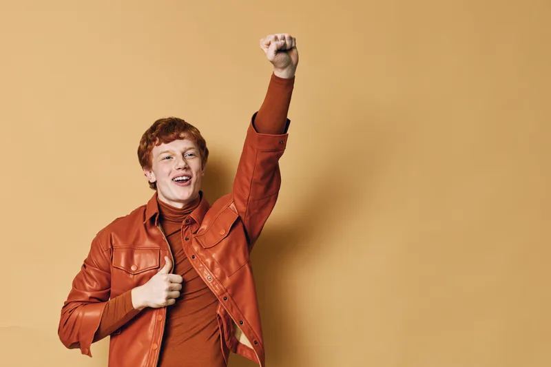 Young man with red hair wearing brown leather jacket and sweater smiling, raising fist and showing thumbs up for success and positive emotions in studio with beige background.