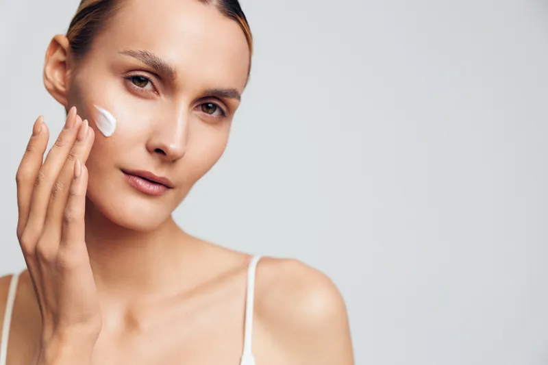 Young woman applying white face cream on her cheek with fingers, close-up portrait on a light background, concept of skincare routine, beauty, and healthy skin care.