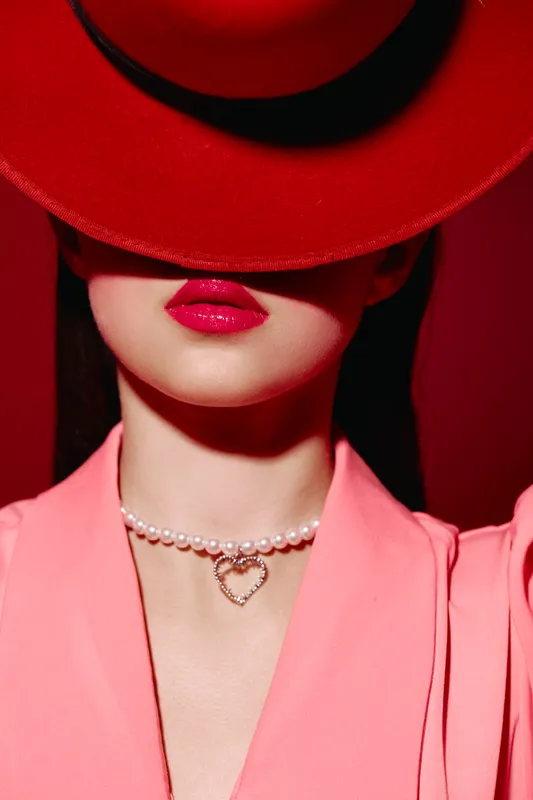 Stylish woman with bold red lips and a large red hat covering eyes wears pink jacket and pearl necklace with heart pendant against vivid red background. Fashion, beauty.