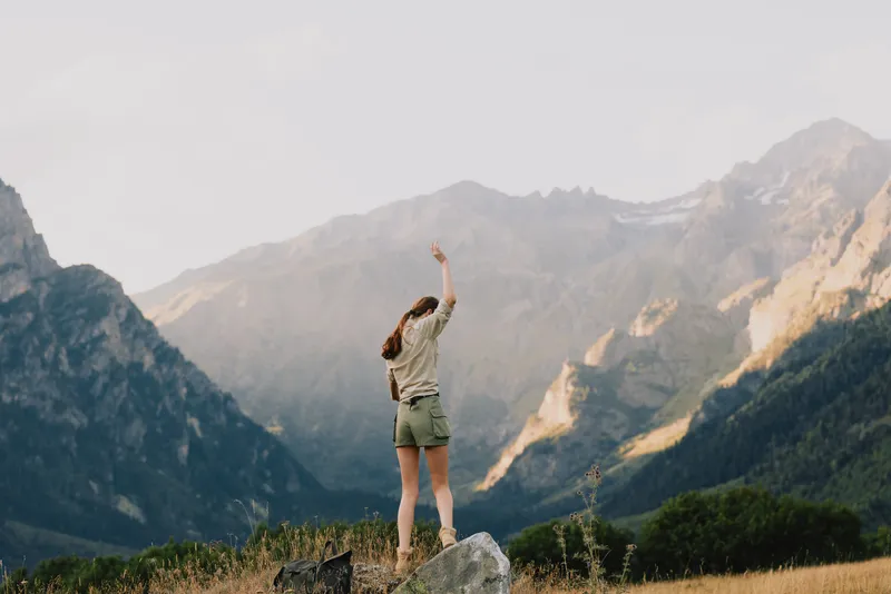 Woman standing on a rock with distant mountains, arms raised. Outdoor lifestyle and nature scene. A moment of freedom, adventure, and wellness in a bright, expansive landscape.