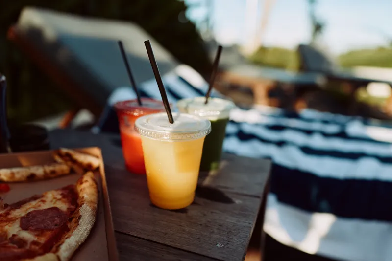 Pizza, drinks, outdoors, casual, summer, snack, meal, relaxation on a wooden table with colorful smoothies beside by a pool, capturing a sunny, shareable lifestyle moment.