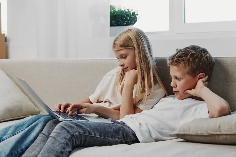 Two children sitting on sofa using laptop together in cozy living room with natural light and relaxed atmosphere.