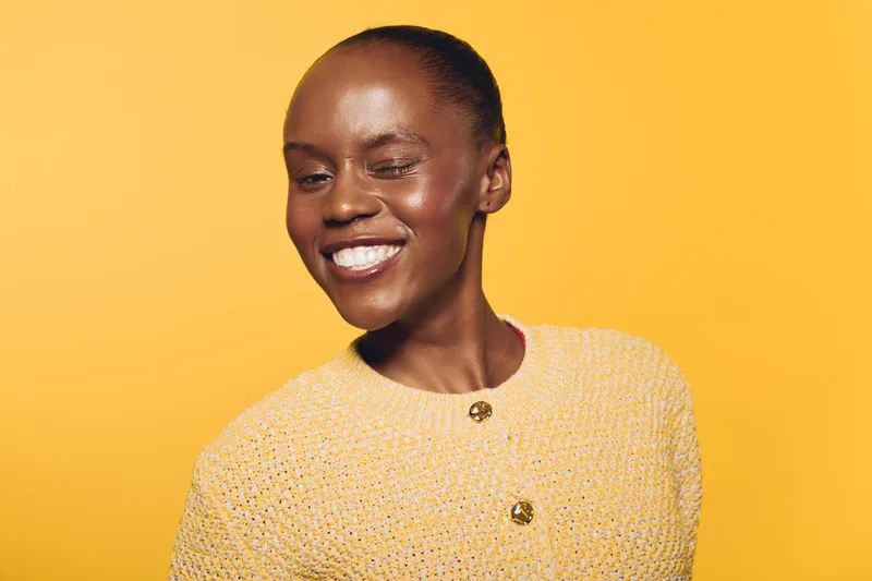 Smiling young African woman with dark skin wearing a yellow knitted sweater winks joyfully against a bright yellow background. Concept of happiness, confidence, and fashion portrait.