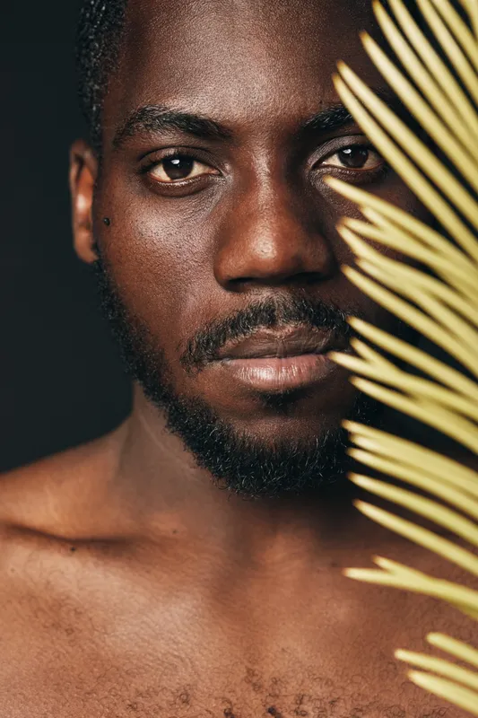 African man with dark skin and beard in creative conceptual visual style holding palm leaf close to face with serious expression on dark background.