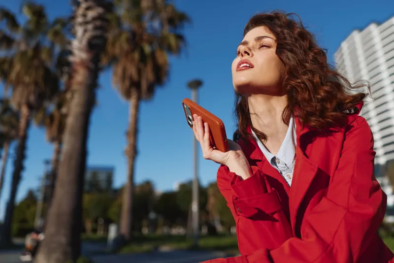 Young woman in a bold red trench coat holds a phone outdoors, standing among palm trees on a sunny street, exuding confidence and a modern urban vibe with bright colors against a blue sky