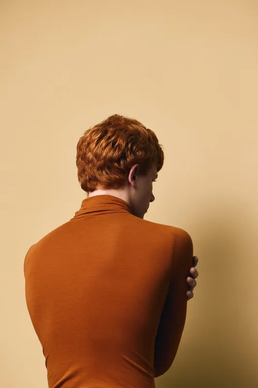 Man with red hair in brown long sleeve shirt seen from behind, standing with crossed arms against beige background in studio setting, casual posture and hairstyle visible.