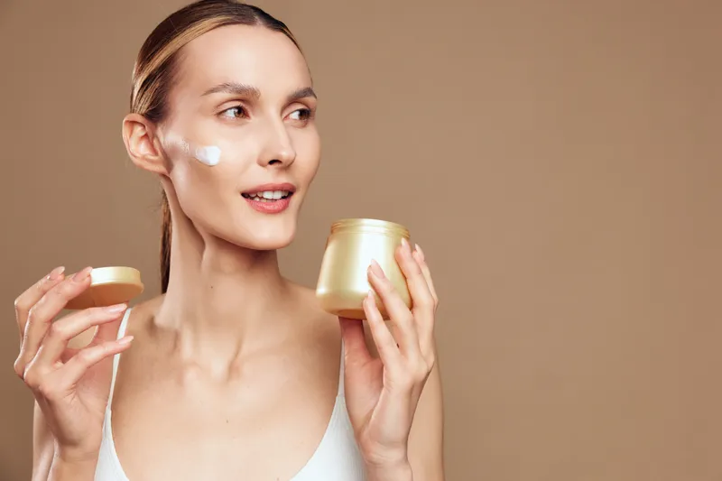 A young woman with clear skin applies face cream on her cheek holding an open jar. She smiles slightly against a plain beige background in a bright studio setting.