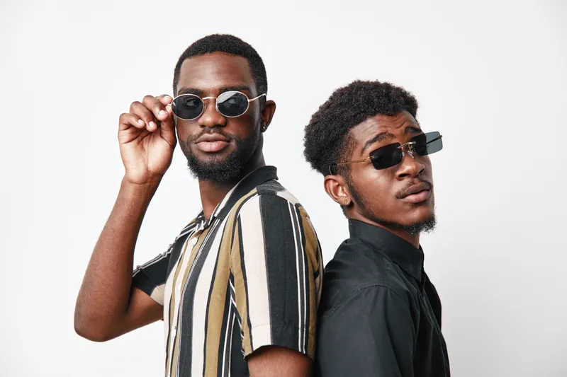 Two african men with dark skin wearing sunglasses and casual shirts posing back to back on white background. Stylish young black males with confident expressions and modern fashion.