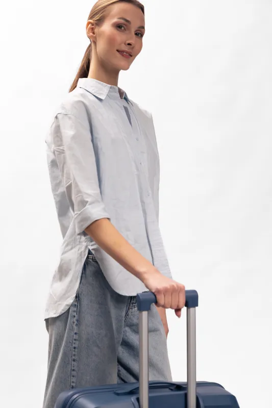 Young woman in casual shirt with suitcase handle, standing on white background. Concept for travel, tourism, and lifestyle with relaxed confident mood and simple modern style.