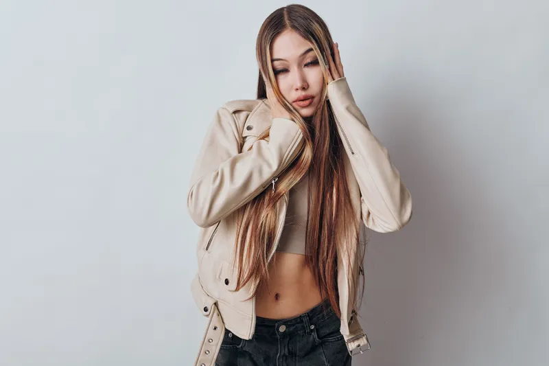 Young woman with long braided hair wearing casual beige jacket and black pants posing with closed eyes on grey background in fashion portrait style indoors.