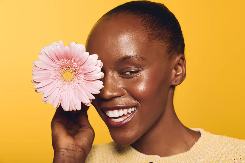 Joyful dark skinned African woman smiles holding pink flower near face against yellow background. Concept of beauty, happiness, skincare, and natural floral wellness in studio portrait.