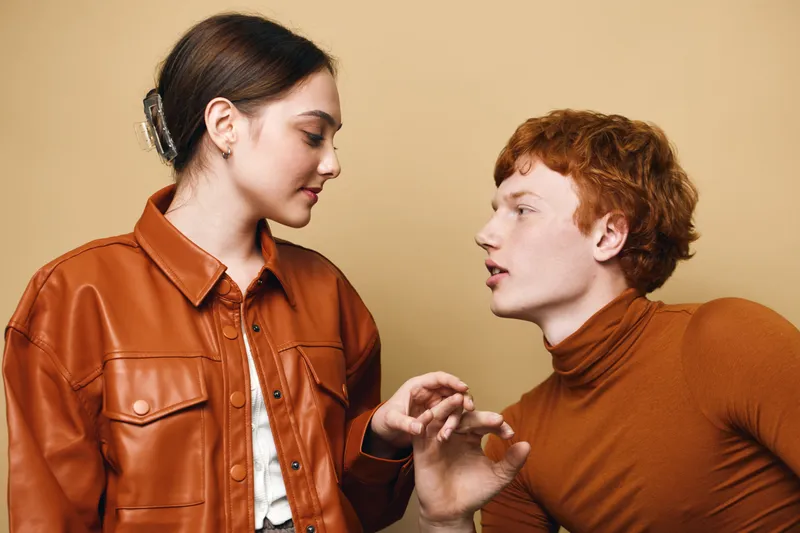 Young man and woman interact with attentive expressions against beige background. Casual clothing includes brown leather jacket and turtleneck sweater in warm tones with natural lighting.