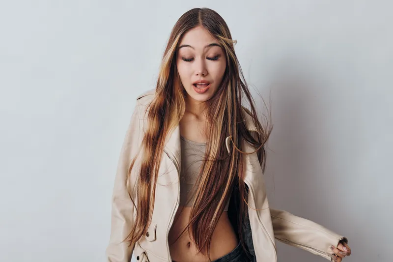 Young woman with long braided hair and beige jacket standing against white background looking down with surprised expression in casual fashion style portrait photography.