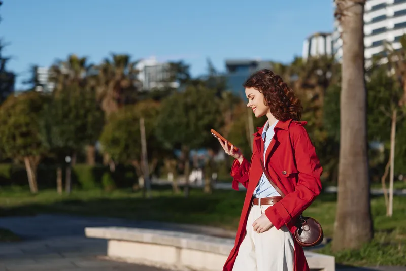 Young woman with curly hair wearing a red coat walks outdoors while looking at her smartphone with a smile in a sunny urban park setting.