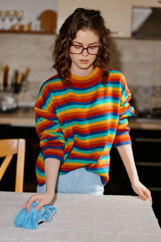 Woman indoors, lifestyle focused home moment, wearing a colorful sweater and glasses, cleaning the kitchen table with a cloth, cozy atmosphere, natural light