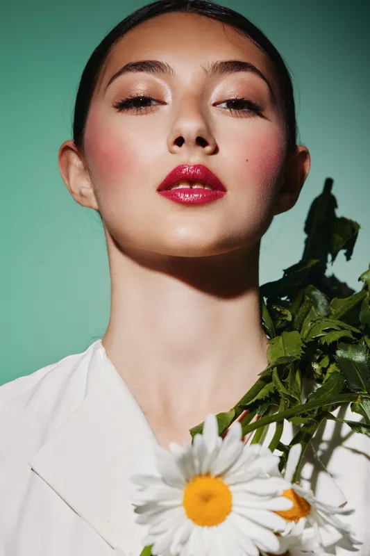 Close-up portrait of a fashionable woman with red glossy lips and dewy skin holding white daisies. Stylish beauty shot with green background for fashion and creative concept.