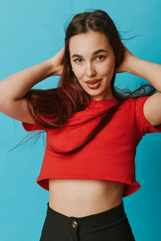 Young woman posing with hands behind head wearing red crop top and black pants against blue background. Female model with long hair and confident expression in casual fashion style.