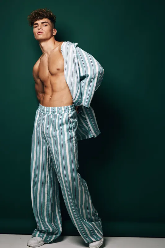 Young muscular man standing with half open shirt and striped pants in studio shooting. Model, male fitness, portrait with curly hair on trendy casual outfit over dark background for fashion.