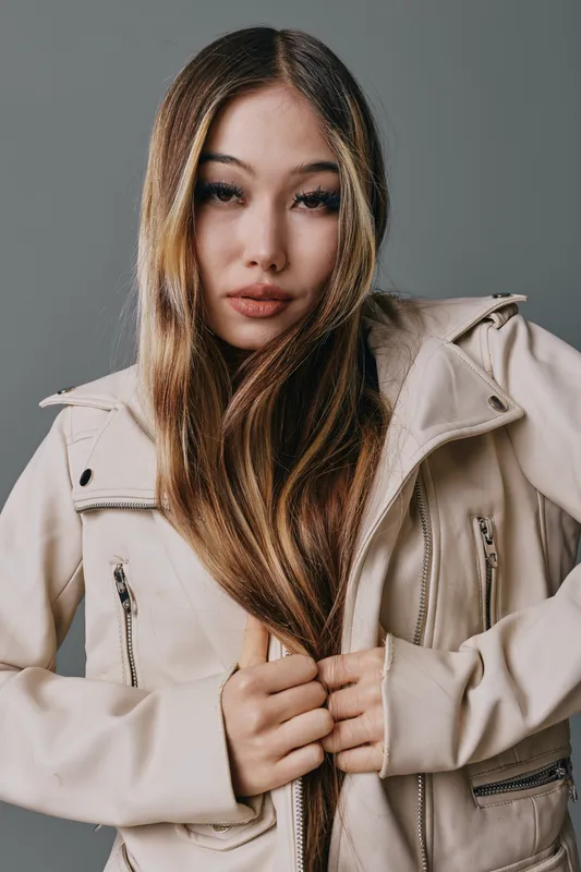 Woman with long hair wearing beige jacket looking at camera, posing with hands holding jacket zipper on gray background. Portrait of female model in casual outerwear fashion style.
