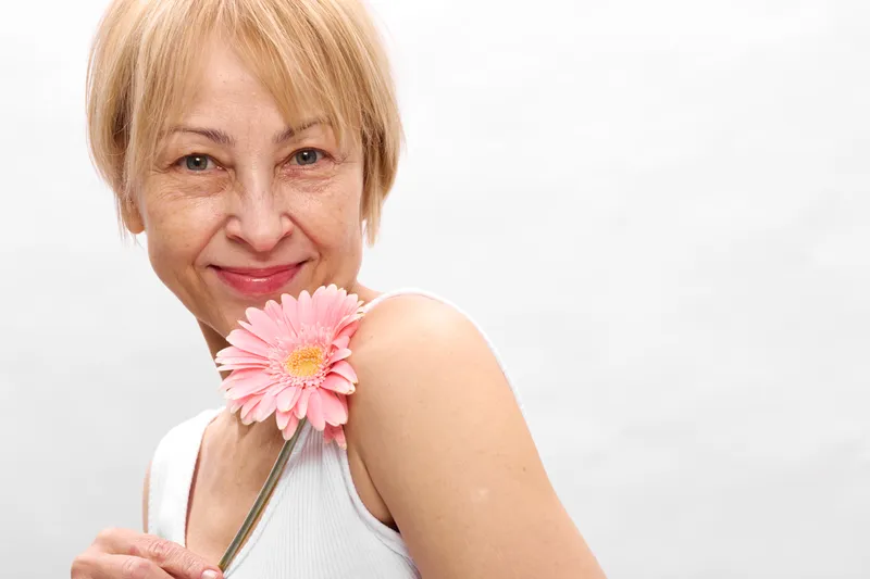 Elderly woman smiling with pink flower, close up portrait in studio, soft natural light, gentle expression, white background, relaxed posture, hand holding stem, serene smile