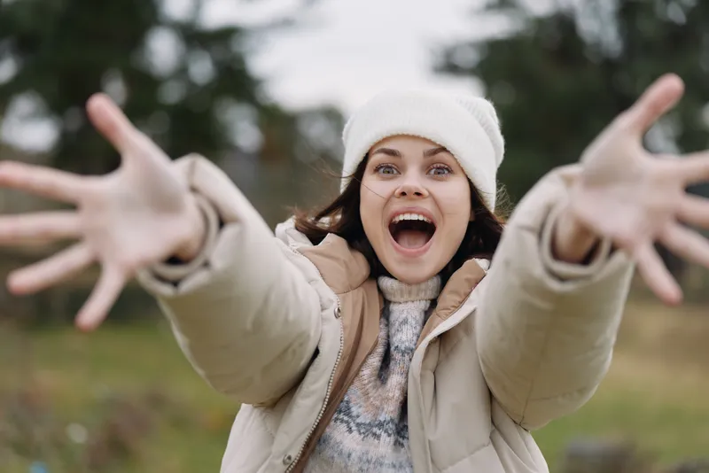 Joyful woman outdoors with open arms, wearing a warm coat and beanie, excited expression toward the camera in a crisp, bright outdoor scene.