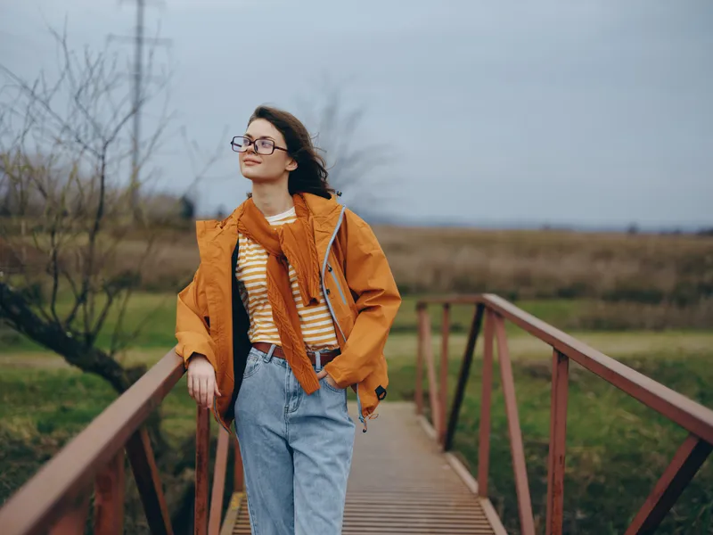 Smiling woman lifestyle outdoors on a wooden bridge, wearing orange jacket and glasses, walking in autumn landscape, cloudy sky soft light, calm nature mood, fresh air experience