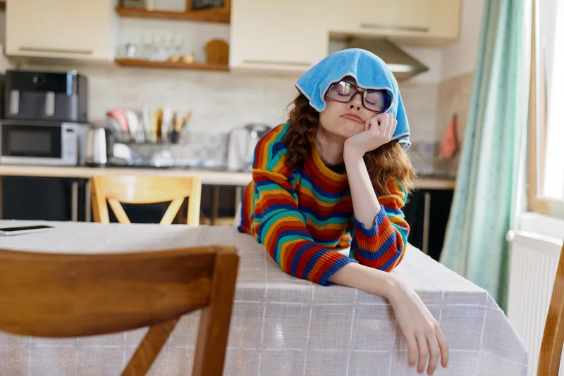 Woman sits at a kitchen table wearing a colorful striped sweater and blue hat, resting her chin on her hand with a thoughtful expression, capturing casual home lifestyle in a cozy indoor scene.